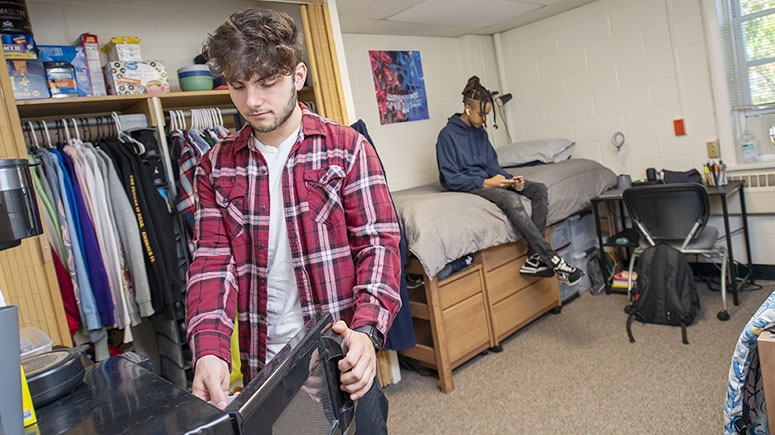 Two students in their room at Richmond Hall: one opens the mini-fridge while the other sits on a bed, engrossed in an electronic device.