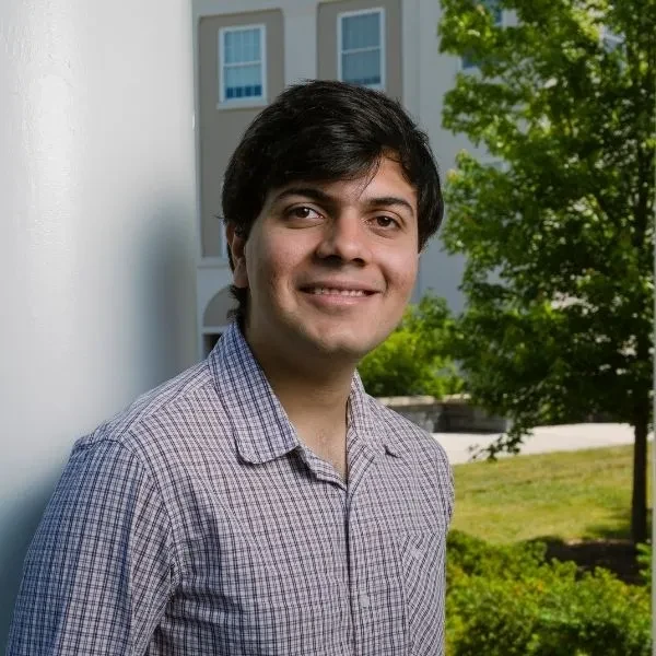 Shams Qureshi ’25 leaning against a column on campus, smiling, with ISEC in the background.