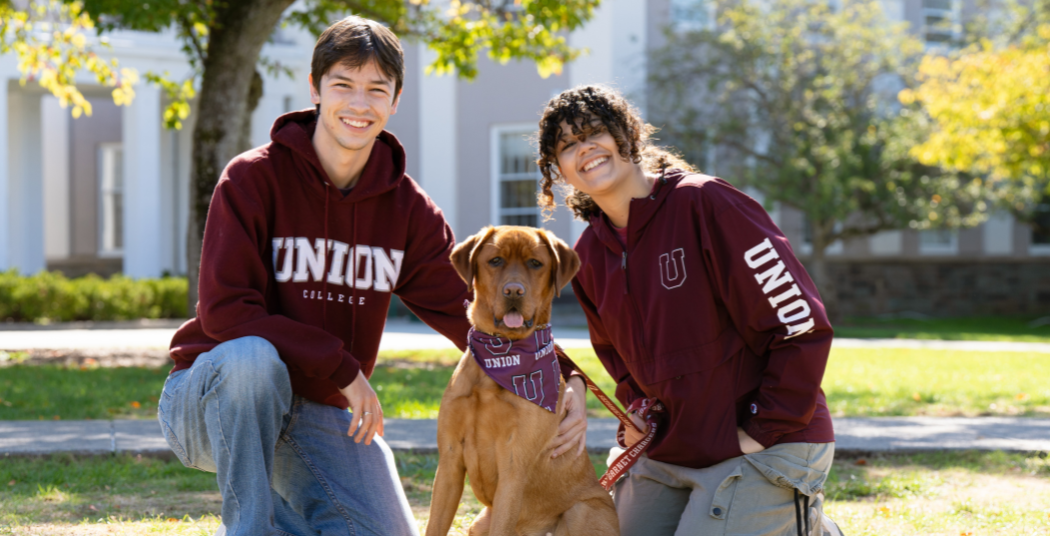 Two students in Union sweatshirts pose with Charger the dog. 