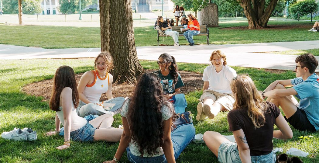 Students sitting outside on campus, chatting with each other on the lawn. 