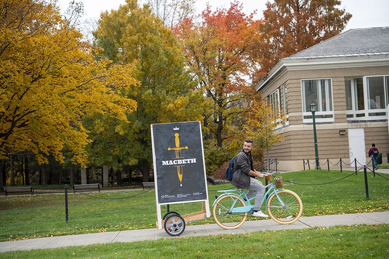 A student on a bike towing a placard advertising an upcoming MacBeth production