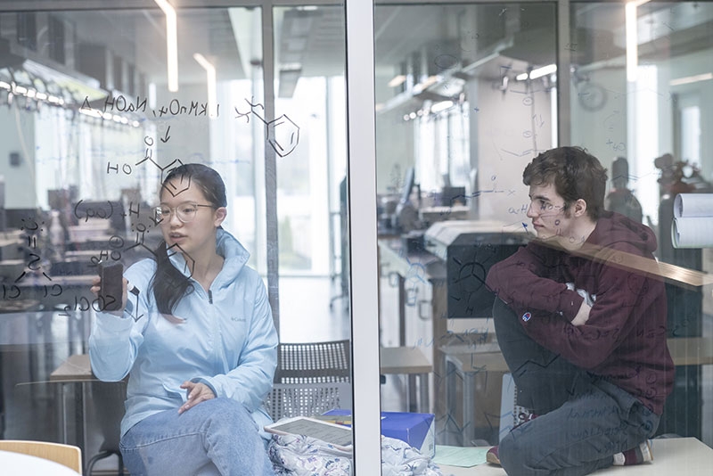 A student writes chemical equations on a partition glass wall in ISEC as another student looks on.