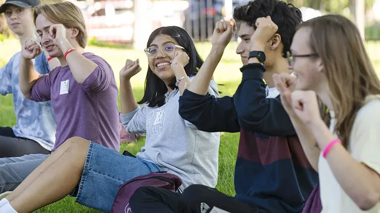 Students participating in group activity that involves raising hands while seated in the grass.
