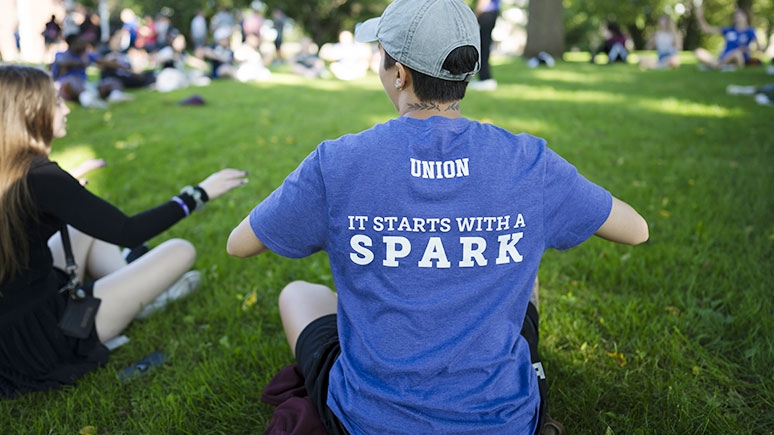 Person sitting on ground wearing a shirt that says it starts with a spark