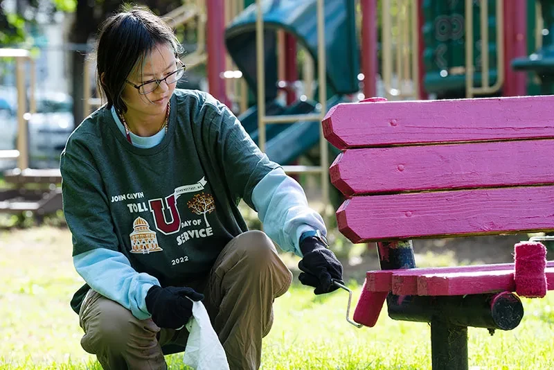 A student participant in Toll Day, with roller in hand, paints a park bench.