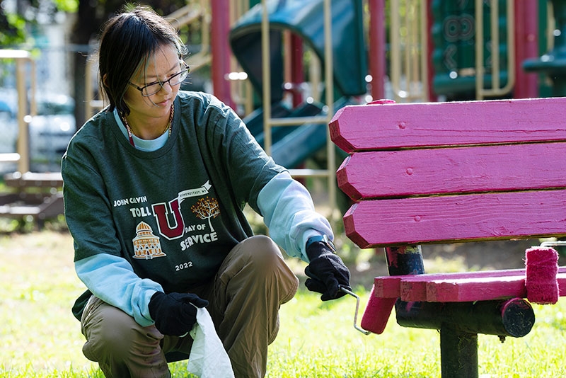 A student participant in Toll Day, with roller in hand, paints a park bench.