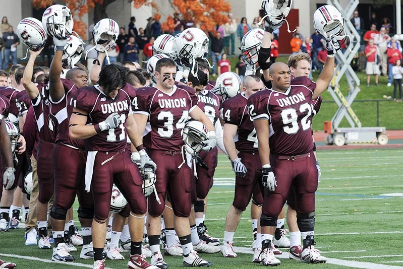  A men's football team standing in formation on the field, with some players raising their helmets in salute.