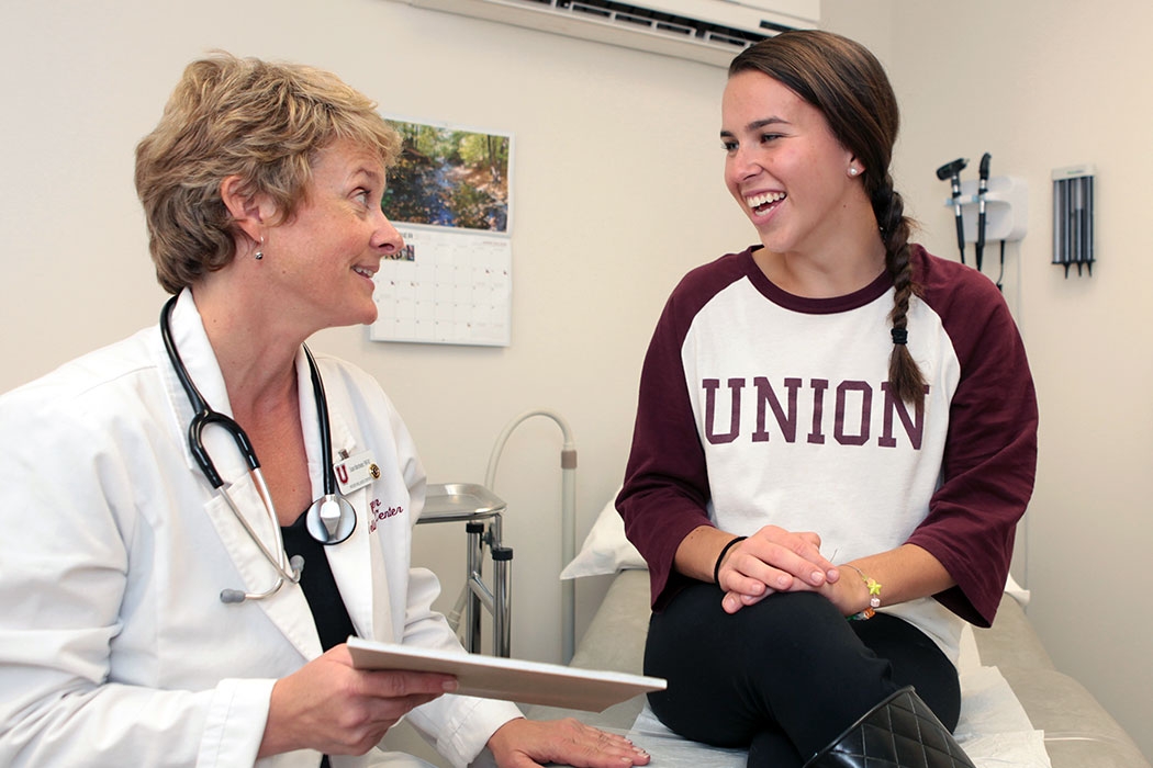 A nurse converses with a student seated on the examination table in an examination room.