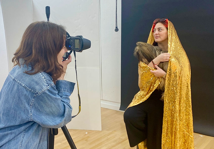 A student poses in a studio with a dog as another student takes their portrait