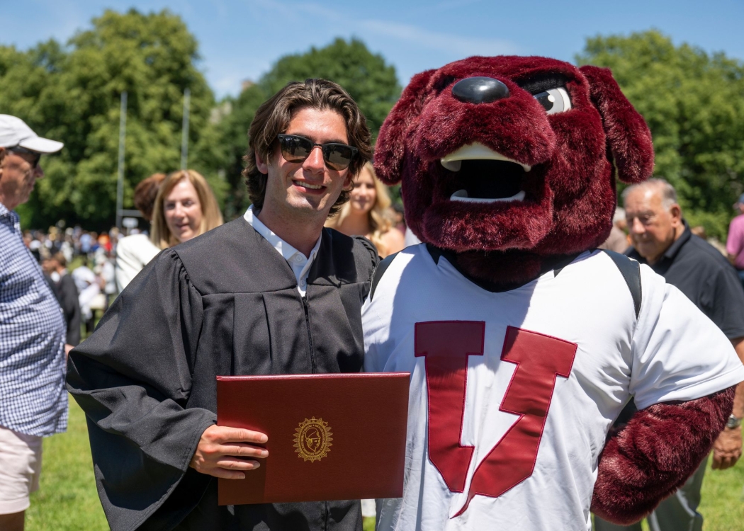 A student at graduation standing next to Charger, proudly holding a diploma case 