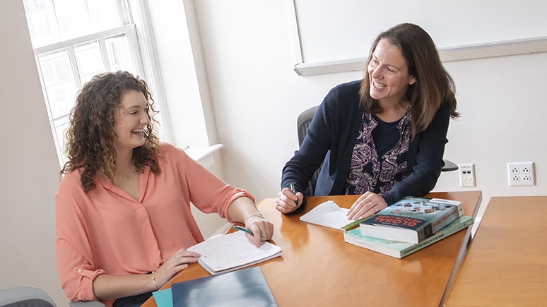 A student and instructor smile as they sit at a table discussing a class assignment