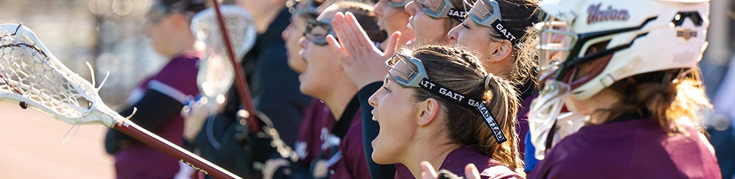 Women's lacrosse players cheer on their teammates from the sidelines.