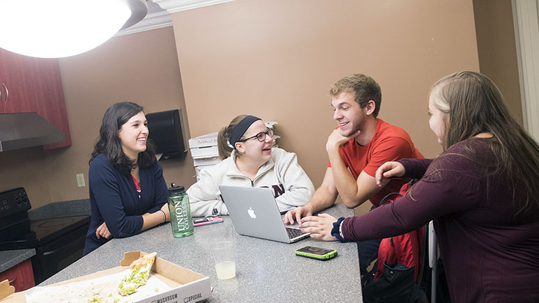 Students gather around a table, laughing and sharing pizza as one student shows something on a laptop.
