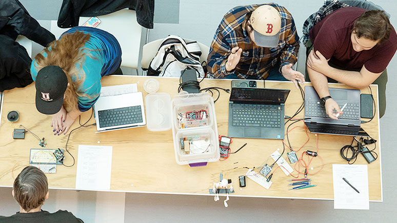 An overhead view of students gathered around a table working on electronic projects, with visible circuits, wires, and other equipment.