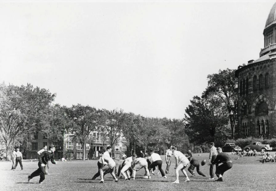 An archival photo of rugby players on the field with the Nott Memorial visible to the side..