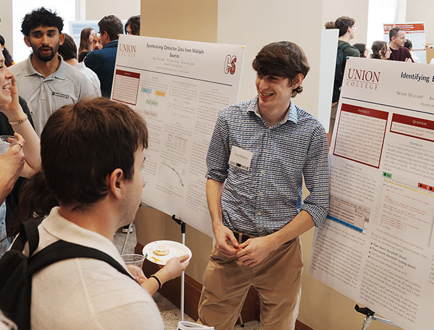 A student stands beside an easel-mounted poster while explaining his research.