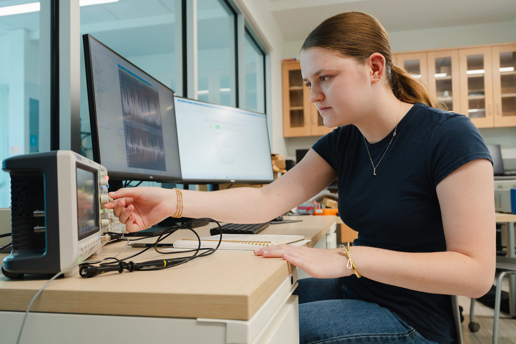 A woman sits at a table in a lab turning the knob on an oscilloscope.