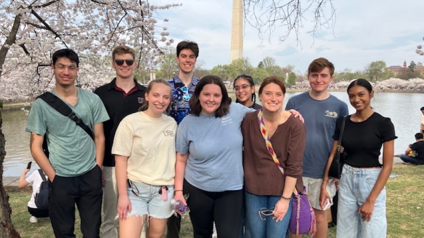 A group of 9 people standing together with the Washington Monument and cherry blossom trees in the background.