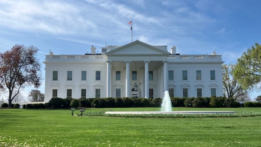 A photo of the White House with the green front lawn and fountain flowing in front of it.