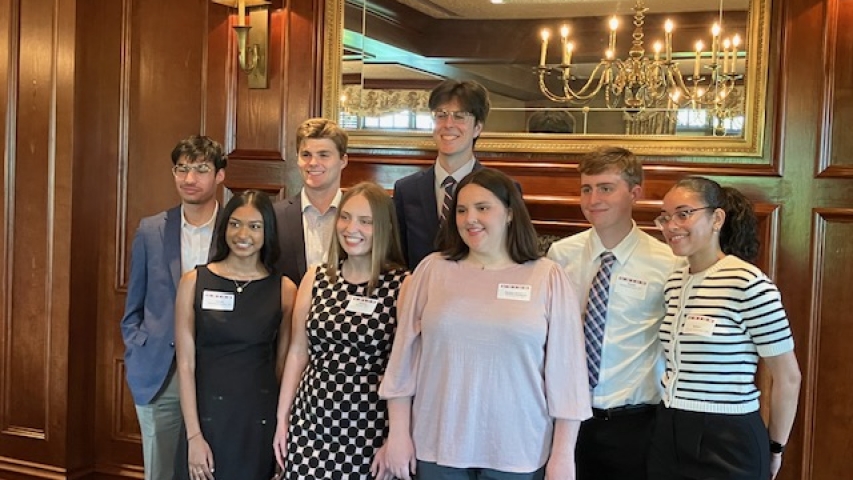 A group of 8 students wearing formal dress standing indoors in front of a dark wooden wall, and a mirror reflecting a lit chandelier.