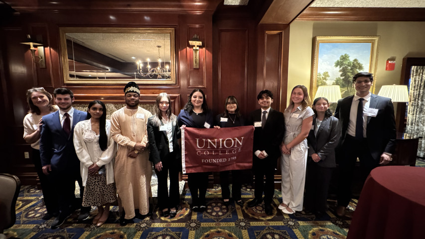 10 students and professor standing with Union college flag in a ballroom