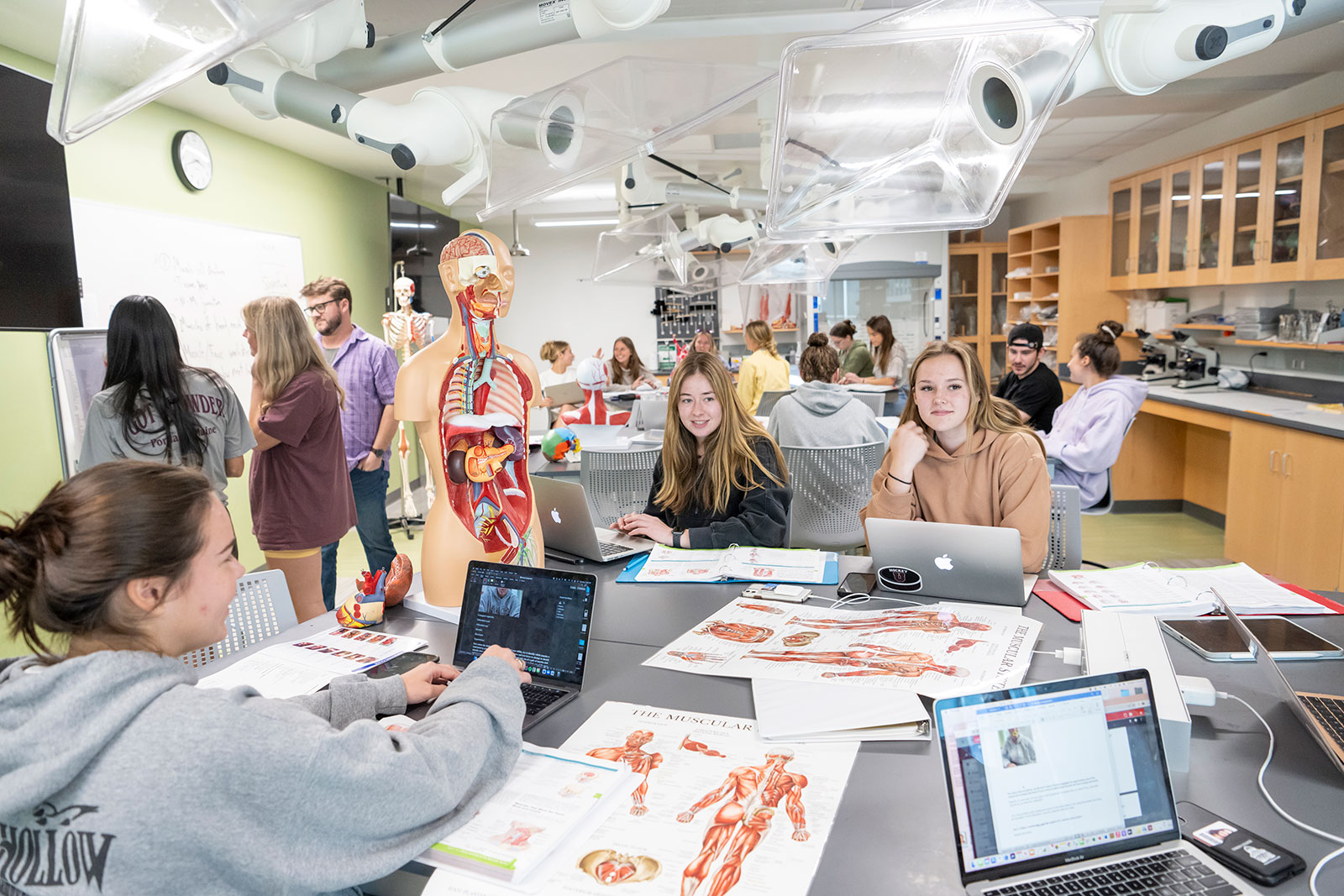 In an anatomy classroom, students are gathered around a table, diligently studying with an anatomical model.