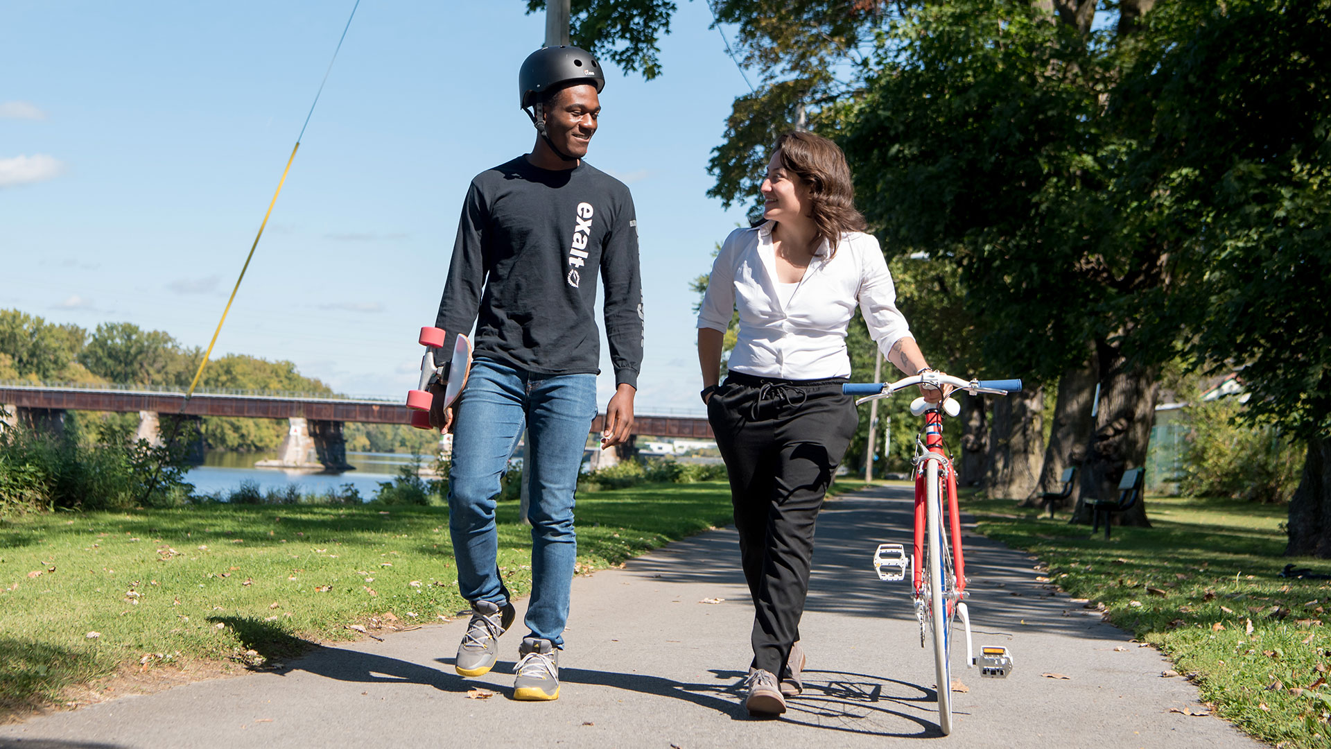 Two students stroll along the Mohawk River bikeway, with one walking her bike and the other holding his skateboard.